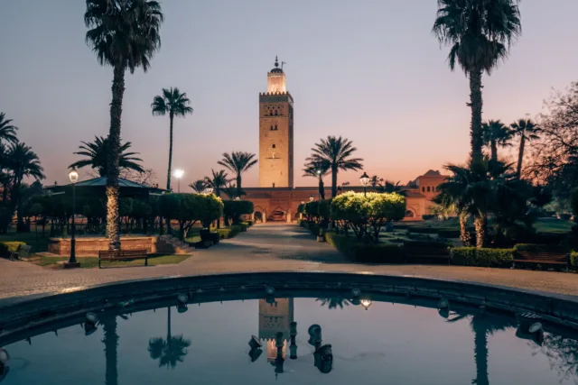 Koutoubia Mosque in Bahja city at sunset, reflected in a still fountain surrounded by palm trees.